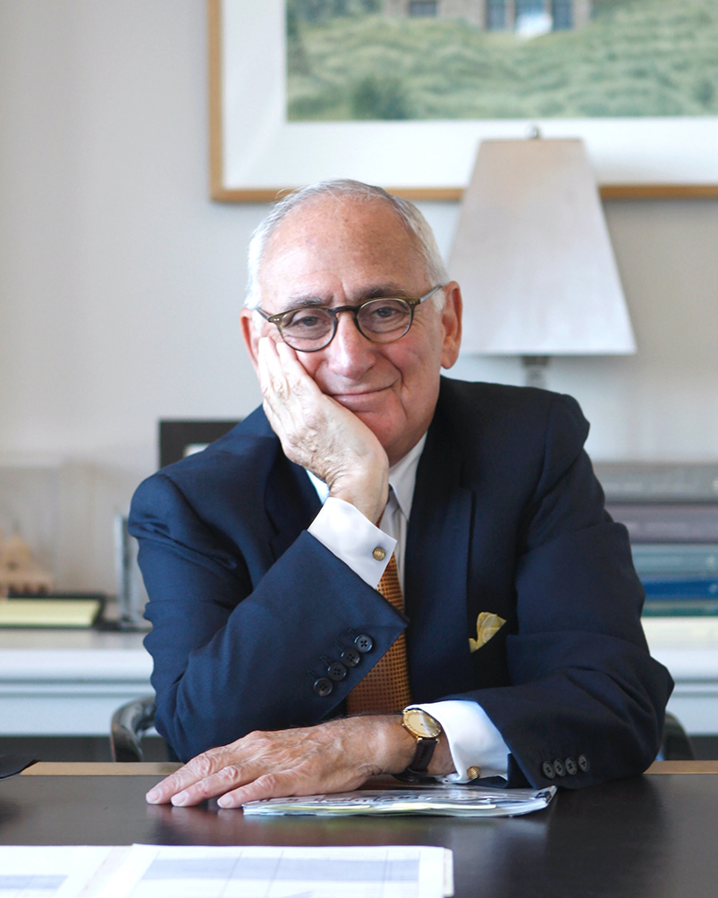 Robert A.M. Stern smiling and sitting on a desk with his hand on his chin